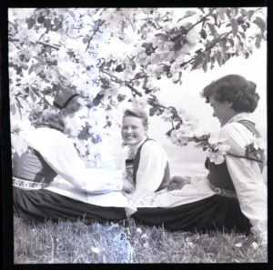 Tre kvinner i bunad, sittande i graset. Foto.